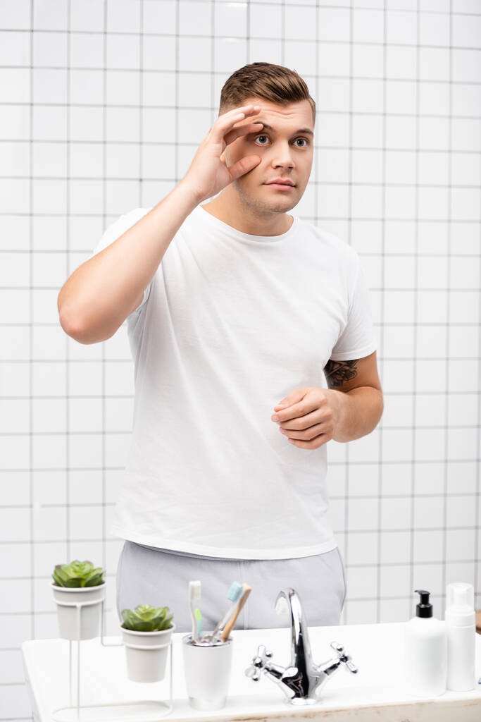 Jeune homme adulte touchant la peau autour de l'œil tout en se tenant près de l'évier dans la salle de bain - Photo, image