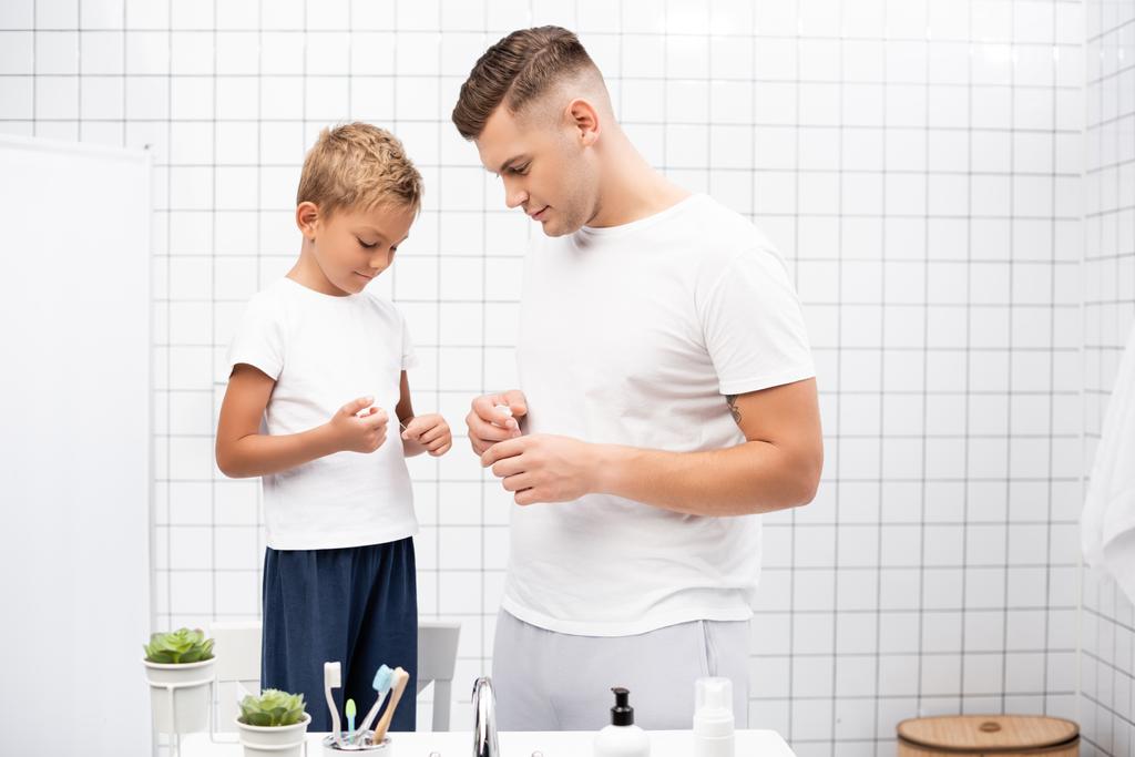 Pozytywny ojciec patrząc na syna trzymającego nitkę dentystyczną stojąc w pobliżu umywalki z kosmetykami w łazience - Zdjęcie, obraz