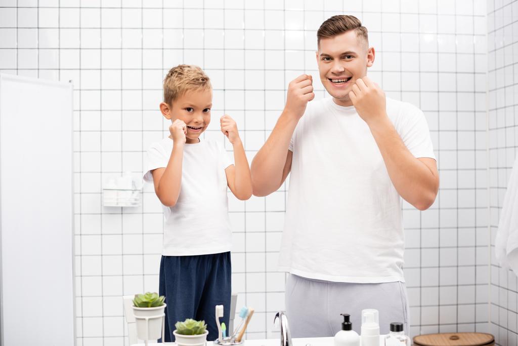 Smiling father and son looking at camera while cleaning teeth with dental floss near sink in bathroom - Photo, Image