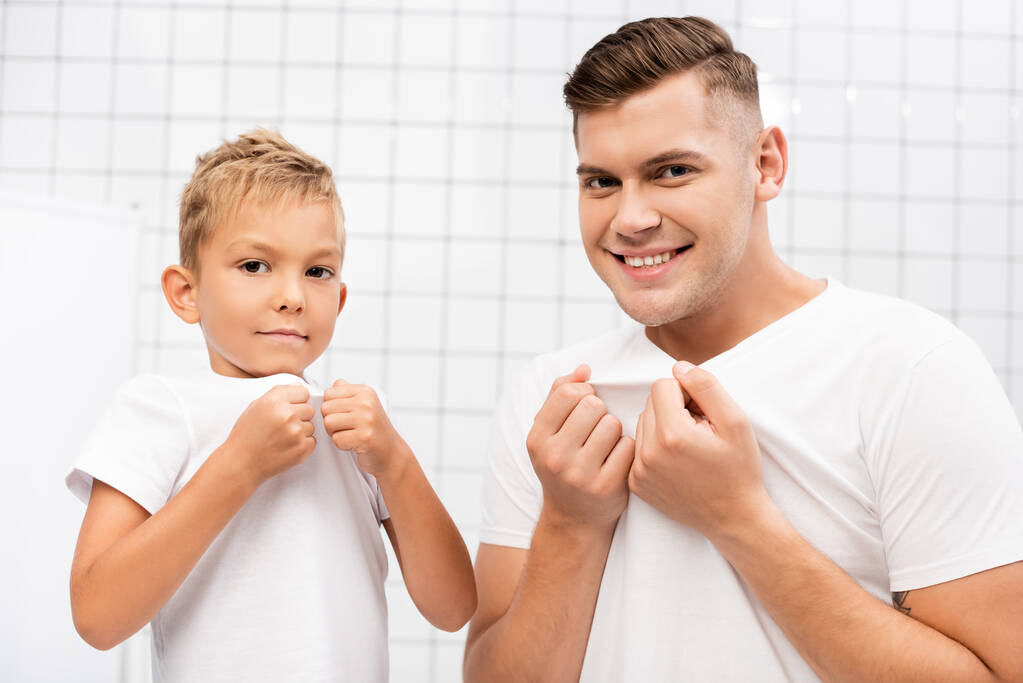 Lächelnder Vater und Sohn ziehen weiße T-Shirts, während sie im Badezimmer in die Kamera schauen - Foto, Bild