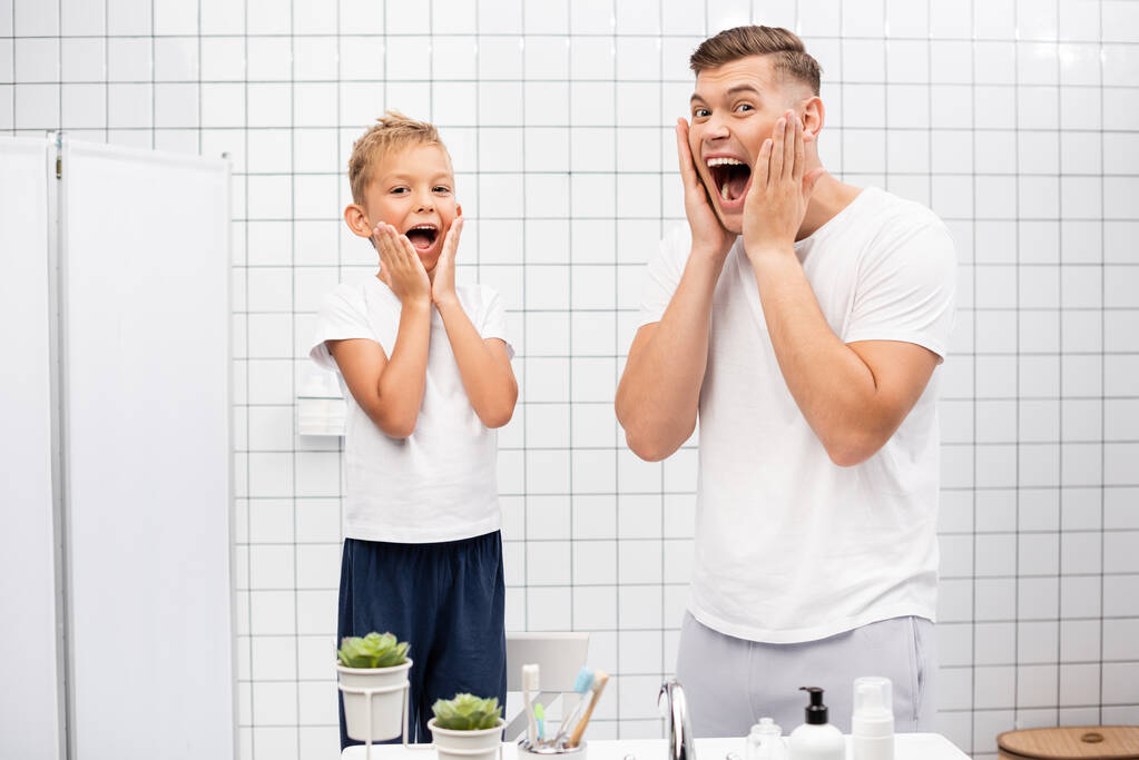 Отец и сын с открытыми ртами касаются щек руками, смотрят в камеру, стоя возле раковины в ванной - Фото, изображение