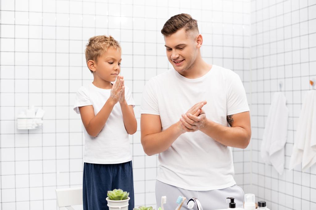Zoon wrijven zeep tussen de handen, terwijl staan op stoel in de buurt van glimlachende vader in de badkamer - Foto, afbeelding