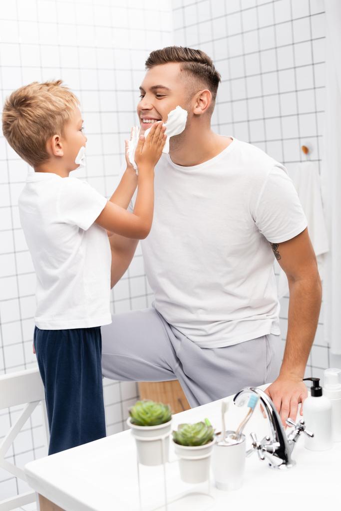 Sohn trägt Rasierschaum auf Gesicht des glücklichen Vaters auf, während er auf Stuhl im Badezimmer steht - Foto, Bild