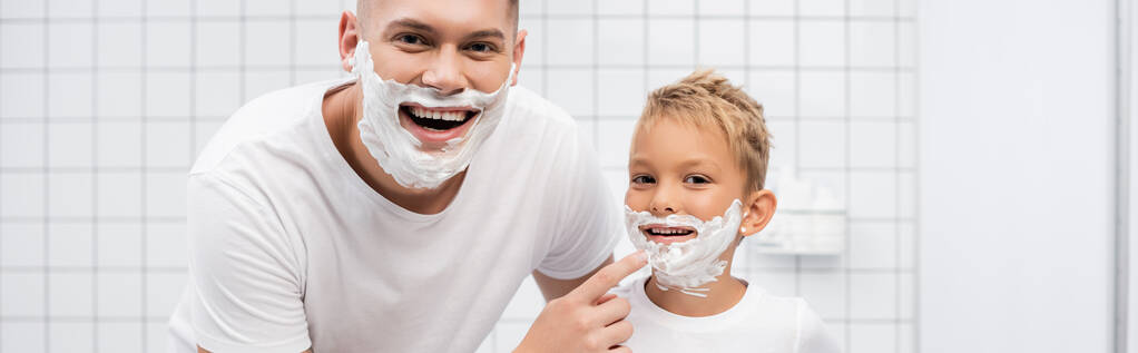 Mutlu baba oğlunun yanında duruyor, yüzünde tıraş köpüğü ve banyoda çenesine dokunuyor, afiş. - Fotoğraf, Görsel