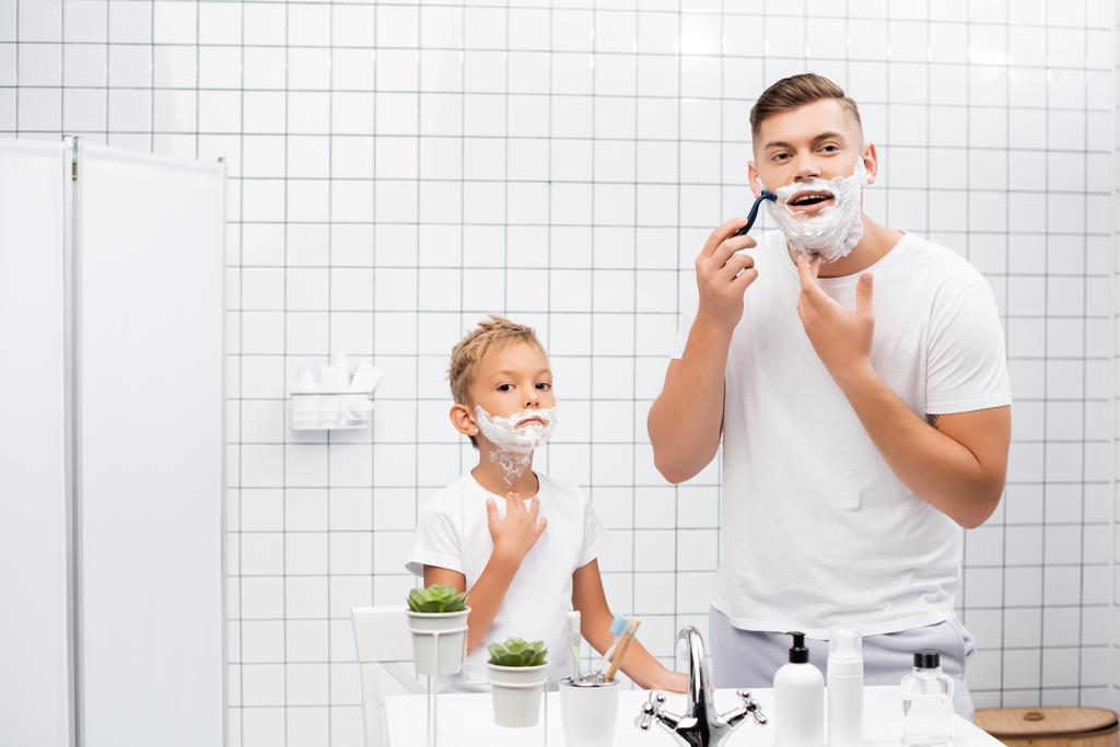 Сосредоточенный мужчина с безопасной бритвой, стоя рядом с мальчиком с пеной для бритья на лице в ванной комнате - Фото, изображение