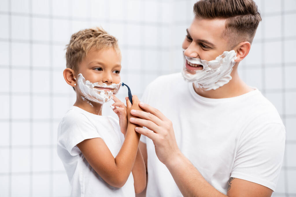 Usmívající se otec při pohledu na syna s pěnou na holení na obličeji drží bezpečnostní holicí strojek u tváře na rozmazaném pozadí - Fotografie, Obrázek