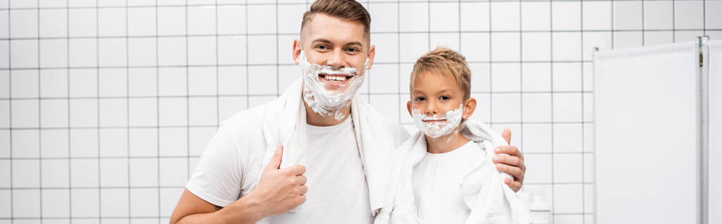 Улыбающиеся отец и сын с пеной для бритья на лицах обнимая и глядя на камеру в ванной комнате, баннер - Фото, изображение