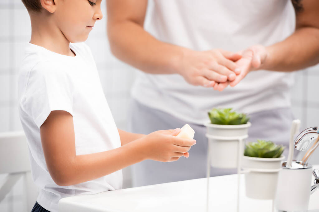Niño preescolar sosteniendo jabón en las manos de pie cerca del fregadero con el hombre borroso en el fondo - Foto, imagen