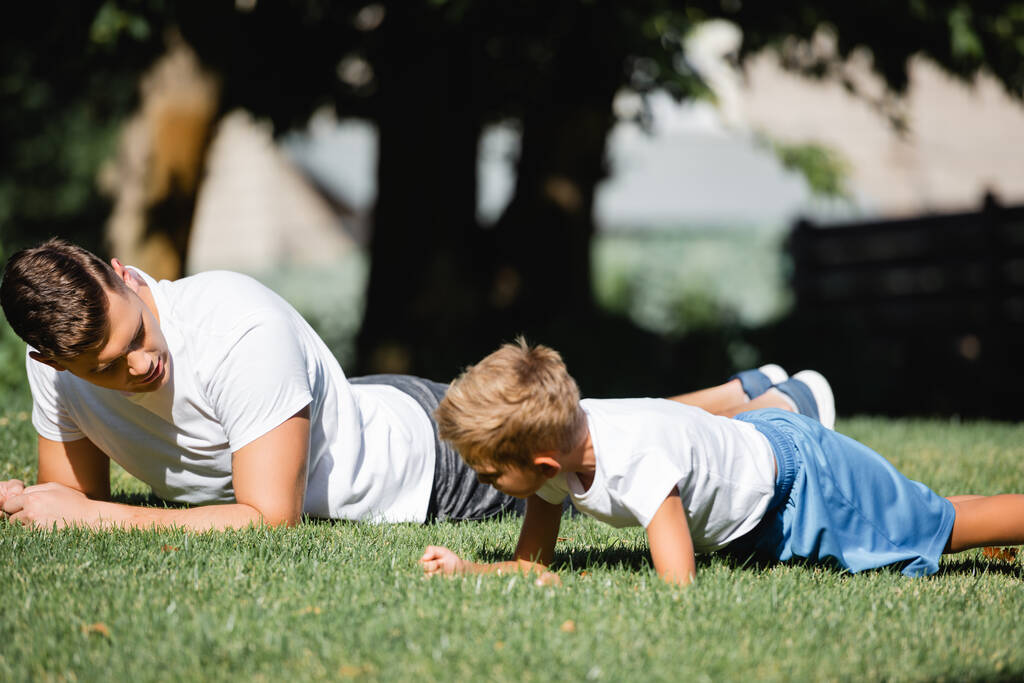 Vader en zoon in sportkleding doen plank op gras in park op wazige achtergrond - Foto, afbeelding