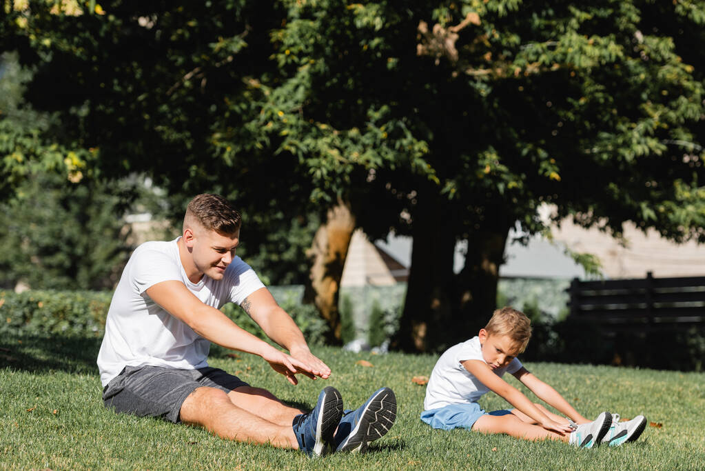 Père et fils en vêtements de sport avec les mains tendues échauffement, tandis que assis sur l'herbe sur fond flou - Photo, image