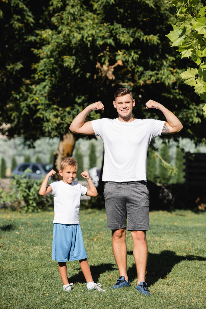 Sonriente padre e hijo en ropa deportiva mirando a la cámara, mientras muestra las manos levantadas con el parque borroso en el fondo - Foto, Imagen