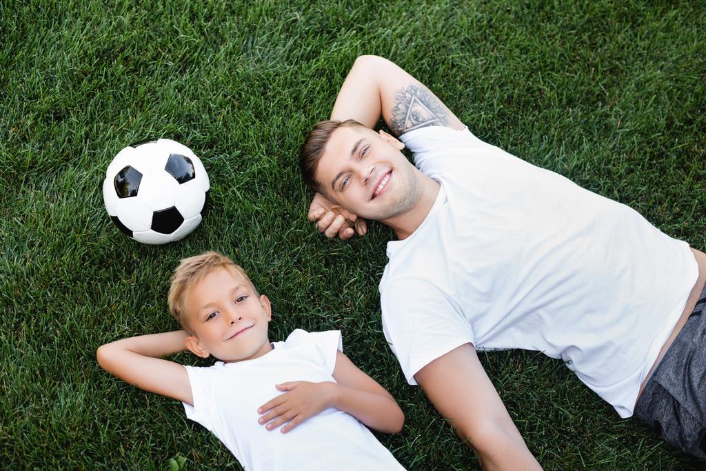 Vista superior de sonriente joven adulto hombre y niño con las manos detrás de las cabezas que yacen cerca de la pelota en la hierba  - Foto, imagen