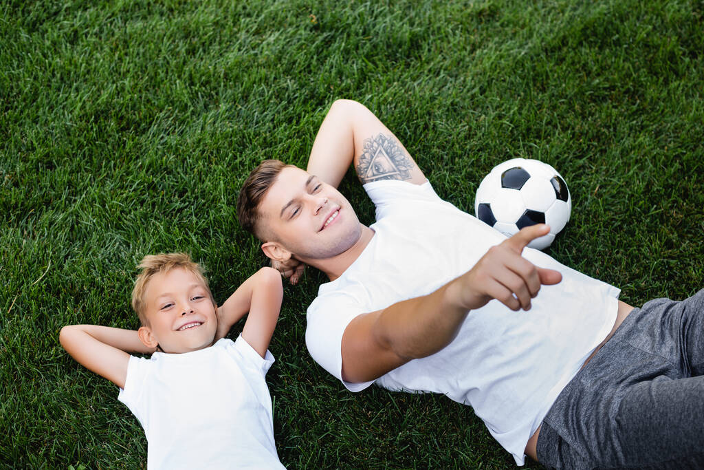Bovenaanzicht van lachende vader wijzend met de vinger, terwijl liggend in de buurt van zoon met handen achter hoofd op gras - Foto, afbeelding