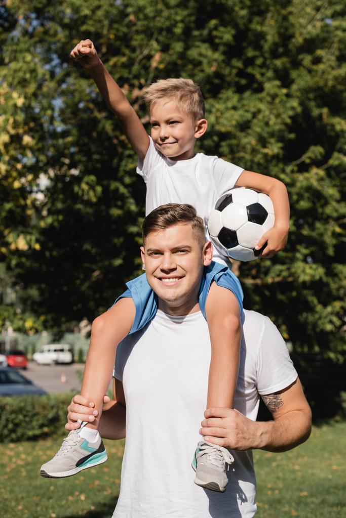 Glücklicher Sohn mit Hand in der Luft, die Ball hält, während er huckepack auf lächelndem Vater im Park auf verschwommenem Hintergrund reitet  - Foto, Bild