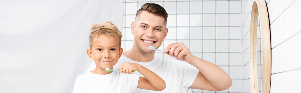 Sonriente hijo y padre sosteniendo cepillos de dientes y mirando a la cámara, pancarta - Foto, imagen