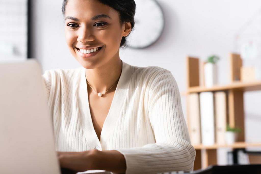 Onnellinen afrikkalainen amerikkalainen naispuolinen työntekijä kirjoittaa kannettavan tietokoneen toimistossa hämärtyneellä etualalla - Valokuva, kuva