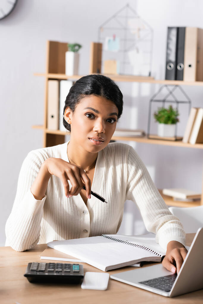 Afroamerikanische Geschäftsfrau mit Stift blickt in die Kamera, während sie auf dem Laptop auf verschwommenem Hintergrund tippt - Foto, Bild