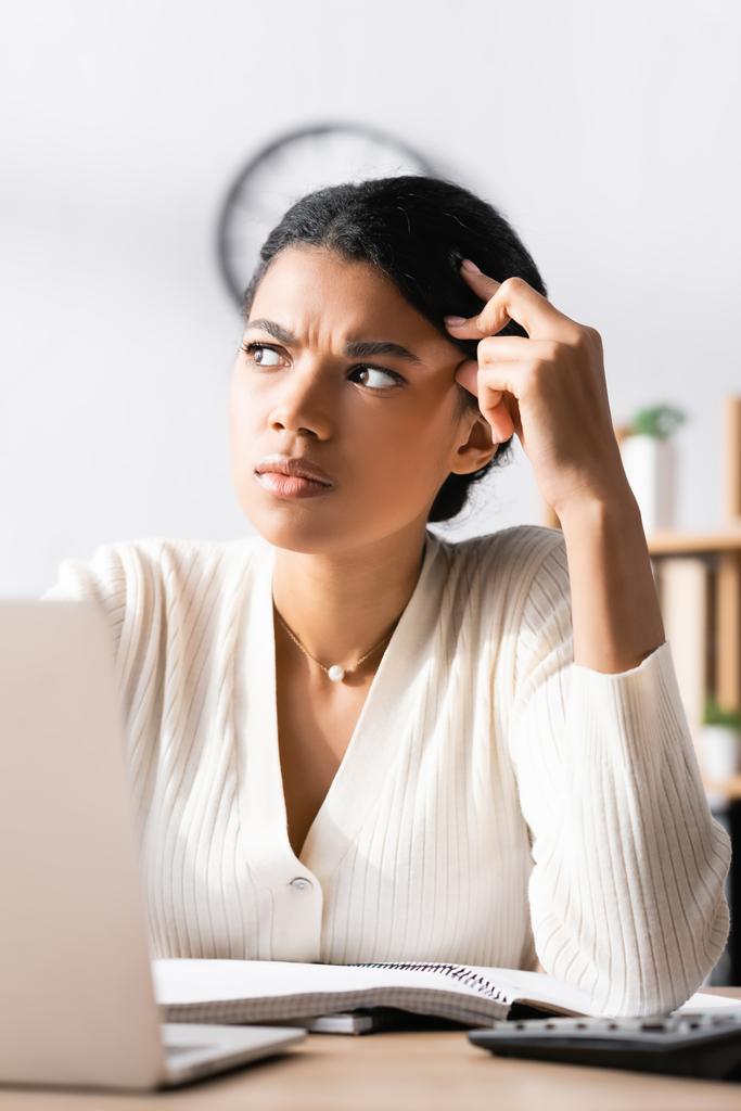 Mujer afroamericana reflexiva mirando hacia otro lado mientras está sentada en el escritorio en la oficina con una computadora portátil borrosa en primer plano - Foto, imagen