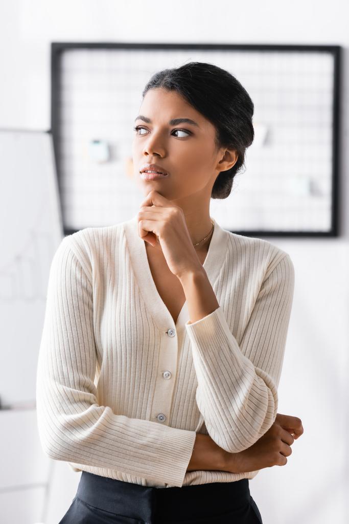 Pensativa mujer afroamericana con la mano cerca de la barbilla mirando hacia otro lado mientras está de pie en la oficina sobre un fondo borroso - Foto, imagen