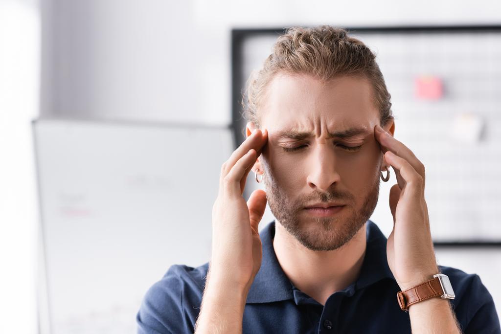 Уставший офисный работник с закрытыми глазами трогает голову руками на рабочем месте на размытом фоне - Фото, изображение