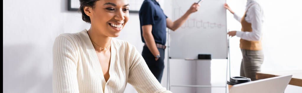 Šťastná africká Američanka při pohledu na notebook na pracovišti s rozmazanými kolegy pracující na pozadí, banner - Fotografie, Obrázek
