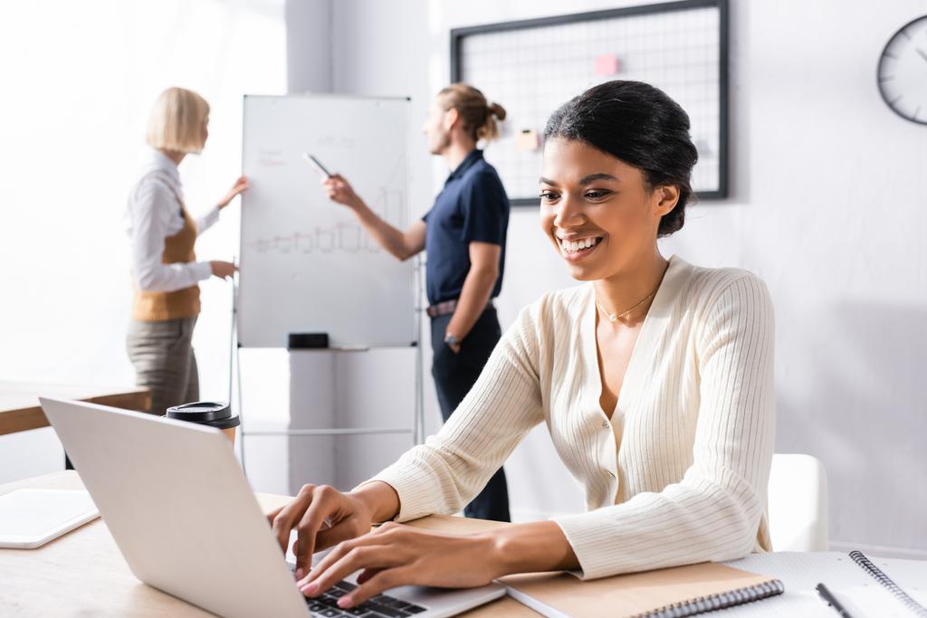 Uśmiechnięta Afroamerykanka pisząca na laptopie siedząc w miejscu pracy z niewyraźnymi kolegami pracującymi nad tłem - Zdjęcie, obraz