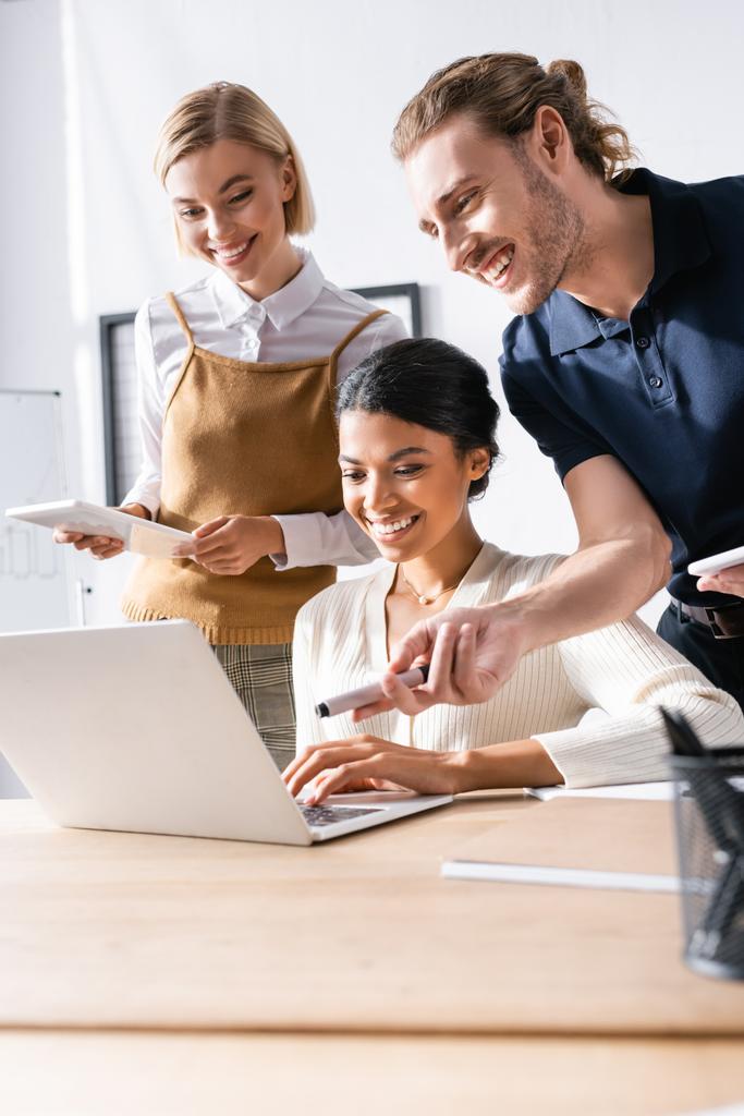 Mann blickt auf Laptop und zeigt mit Filzstift, während er neben multikulturellen Büroangestellten am Arbeitsplatz steht - Foto, Bild