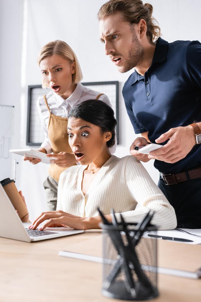 Geschokte multiculturele kantoormedewerkers op zoek naar laptop op de werkplek met wazige pennenhouder op de voorgrond - Foto, afbeelding