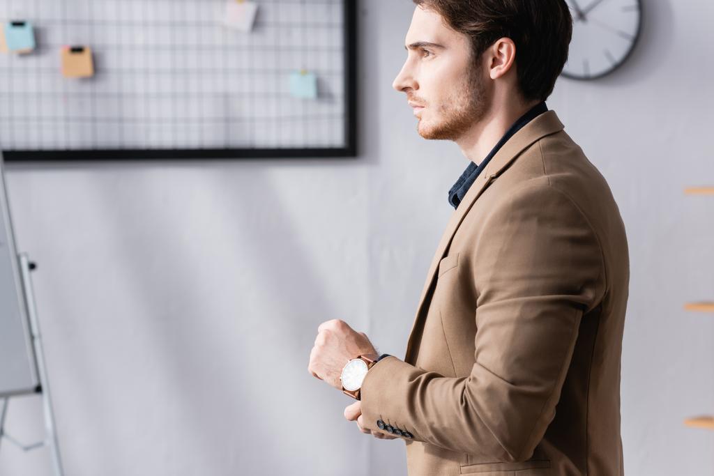 Вид сбоку уверенный бизнесмен стоит и смотрит в сторону, в то время как выпрямить рукав пиджака в офисе на размытом фоне - Фото, изображение
