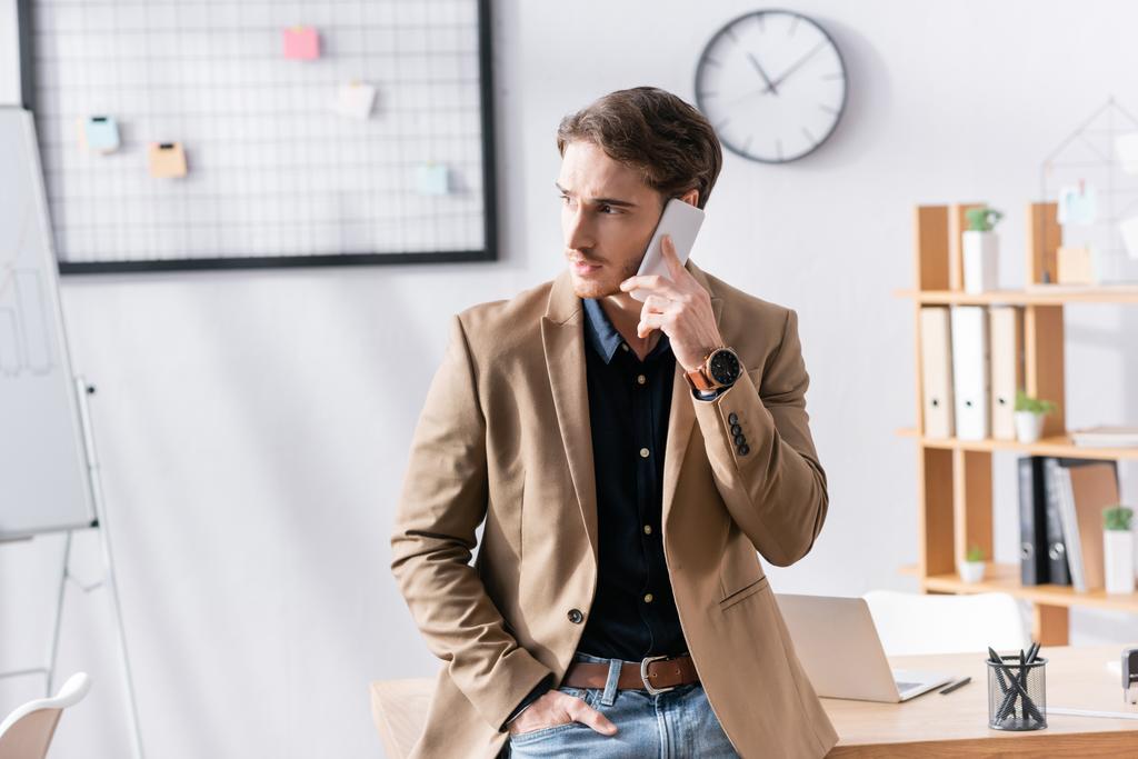 Itsevarma mies, jolla on käsi taskussa, joka katsoo poispäin puhuessaan puhelimessa, nojaa toimistossa toimistossa hämärtyneeseen taustaan - Valokuva, kuva