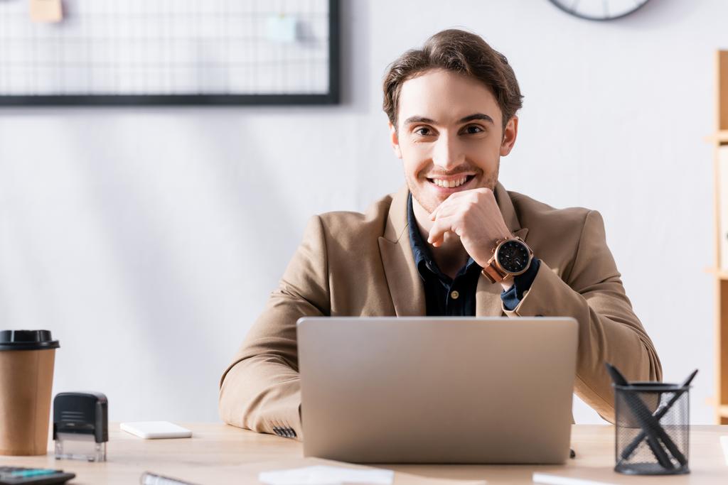 Glimlachende kantoormedewerker die naar de camera kijkt terwijl hij in de buurt van een laptop op kantoor zit op een wazige achtergrond - Foto, afbeelding