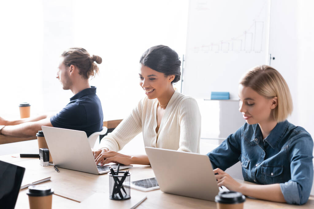 Mosolygó multikulturális női munkatársak gépelnek laptopon, miközben irodai kollégájuk mellett ülnek az asztalnál - Fotó, kép