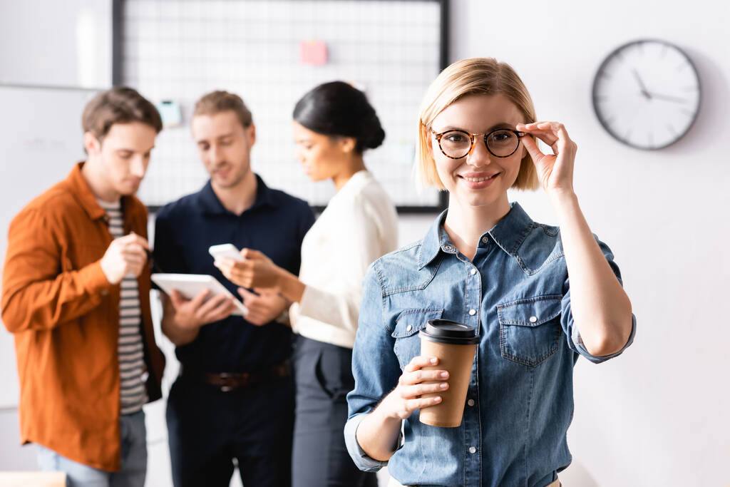 sonriente gerente tocando anteojos y sosteniendo café para ir mientras colegas multiétnicos discutiendo sobre fondo borroso - Foto, imagen