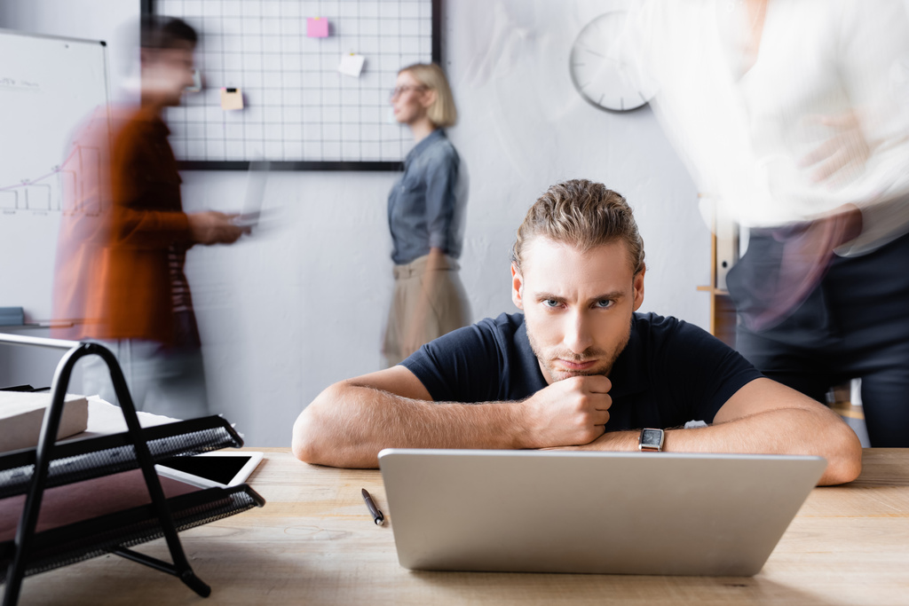 ernsthafter Manager sitzt mit dem Kopf auf der Hand in der Nähe von Laptop im Großraumbüro, Bewegungsunschärfe - Foto, Bild