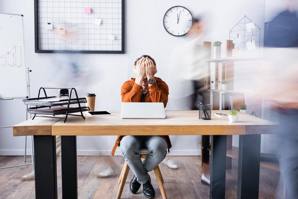 wyczerpany biznesmen zakrywający twarz dłońmi podczas siedzenia w miejscu pracy w biurze otwartej przestrzeni, baner, rozmycie ruchu - Zdjęcie, obraz