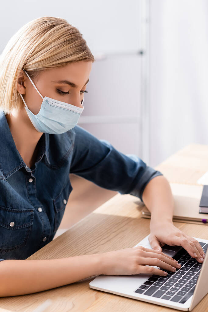 молодой менеджер в медицинской маске, печатающий на ноутбуке в офисе - Фото, изображение