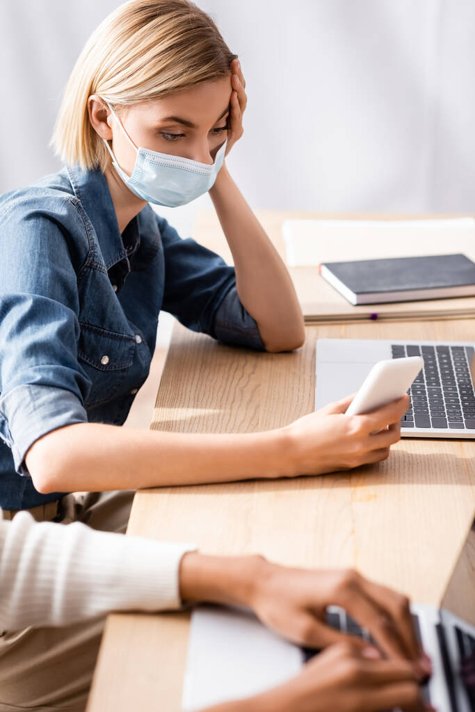 Blonde Geschäftsfrau in medizinischer Maske mit Smartphone in der Nähe eines amerikanischen Kollegen - Foto, Bild