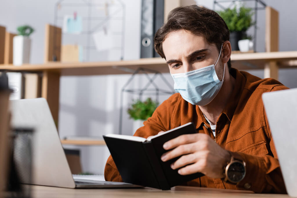 νεαρός επιχειρηματίας με ιατρική μάσκα κοιτάζοντας στο σημειωματάριο στο χώρο εργασίας σε θολή φόντο - Φωτογραφία, εικόνα