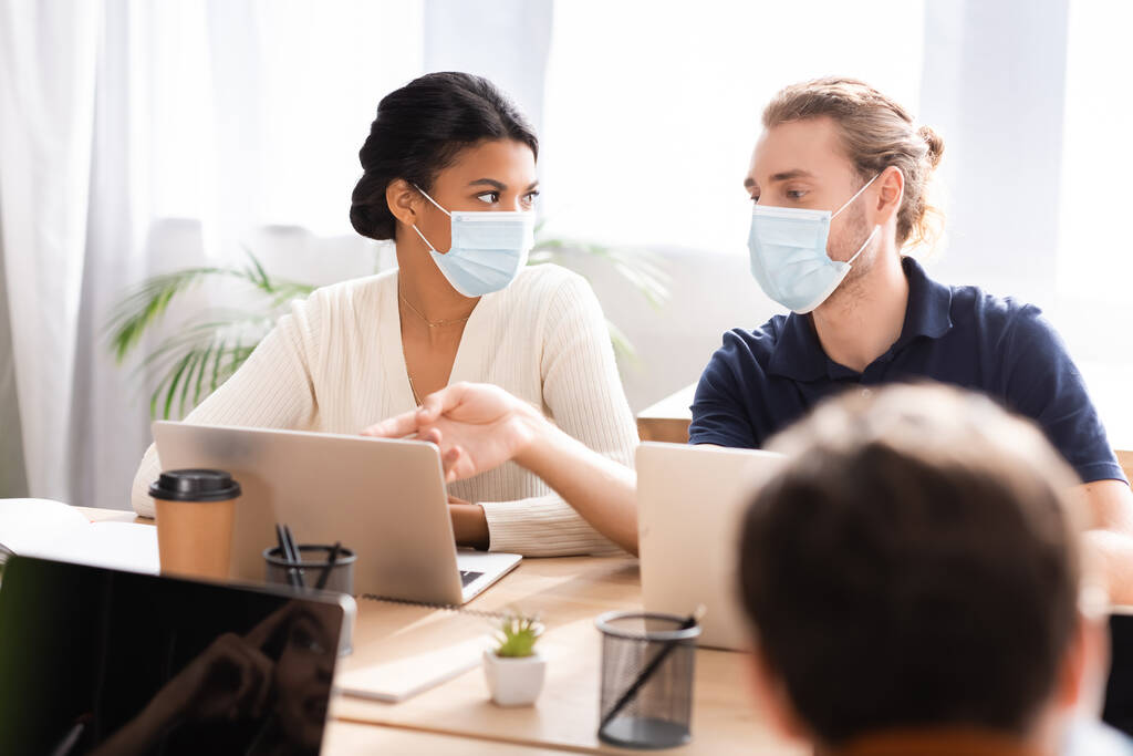 manager in medical mask pointing with finger at laptop near african american colleague, blurred foreground - Photo, Image