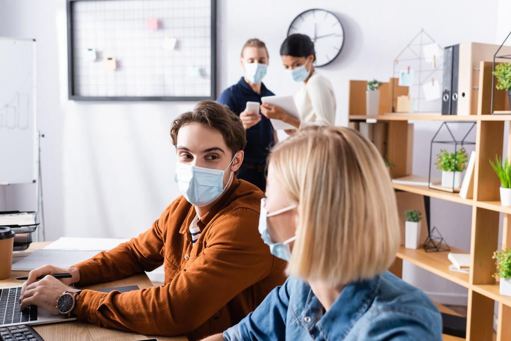 молодой менеджер в медицинской маске смотрит на коллегу, в то время как бизнесмены разговаривают на размытом фоне - Фото, изображение