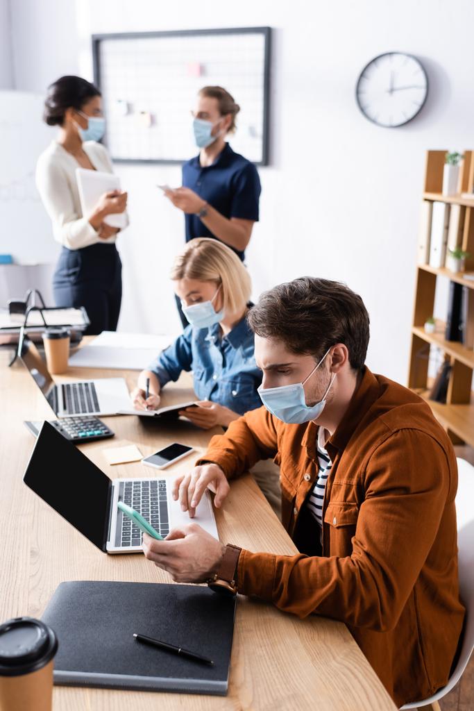 mladí multikulturní podnikatelé v lékařských maskách pracující v otevřeném prostoru kanceláře během karantény na rozmazaném pozadí - Fotografie, Obrázek
