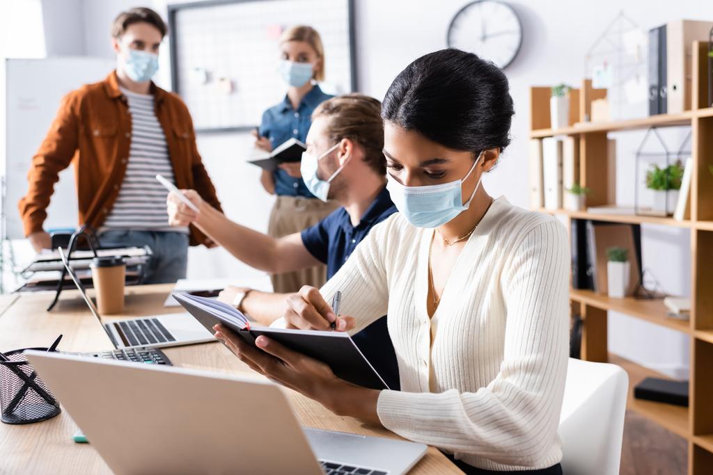 africký americký podnikatelek v lékařské masky psaní v notebooku v blízkosti kolegové pracující na rozmazaném pozadí - Fotografie, Obrázek