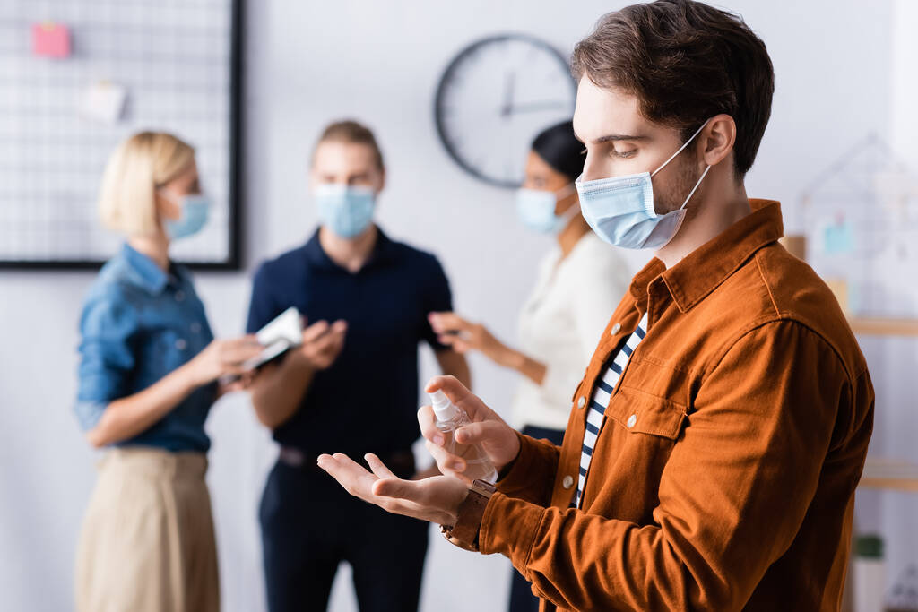 gerente em máscara médica pulverização desinfetante em mãos perto de colegas em fundo turvo - Foto, Imagem