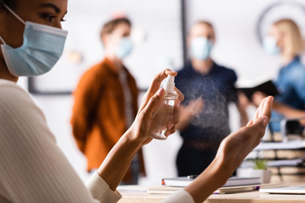 partial view of african american manager disinfecting hands at workplace, and colleagues  standing on blurred background - Photo, Image