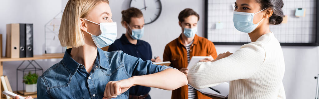 nuoret liikenaiset lääketieteellisissä naamioissa tekemässä kyynärpää kolahtaa lähellä kollegoita hämärtyneellä taustalla, banneri - Valokuva, kuva