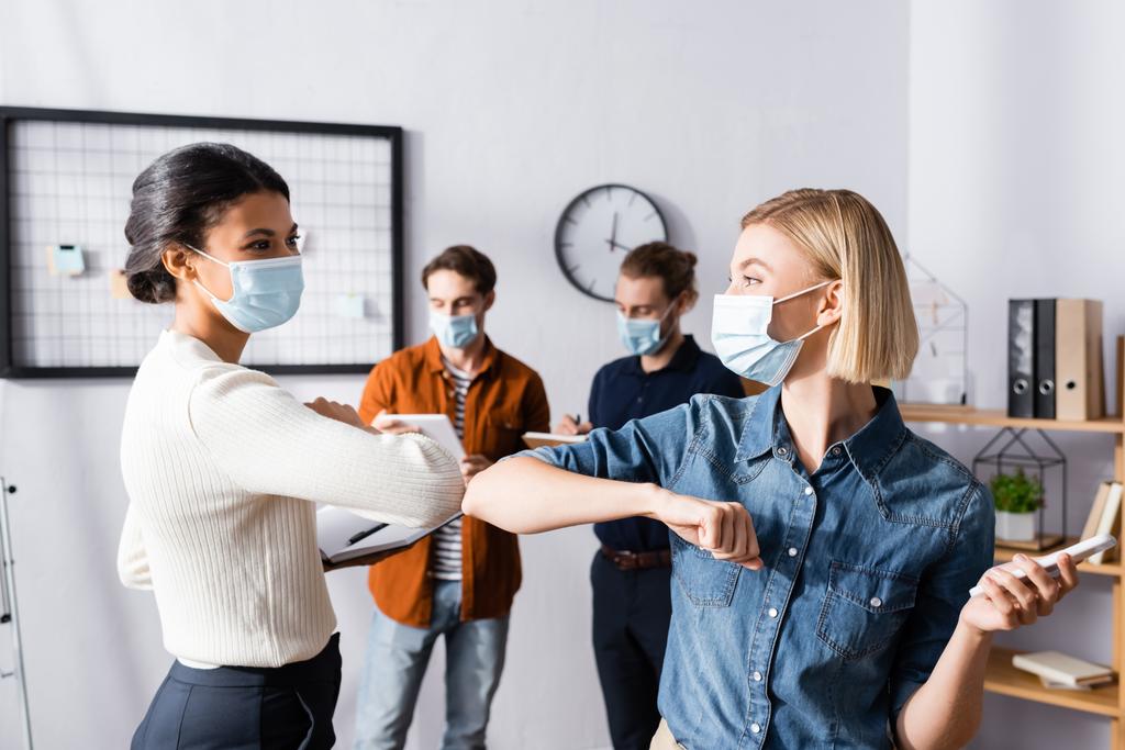 jeunes gestionnaires dans des masques médicaux faisant geste de bosse du coude près des hommes d'affaires sur fond flou - Photo, image