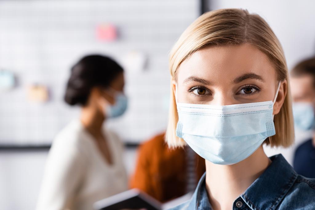 blond zakenvrouw in medisch masker kijken naar camera terwijl multi-etnische collega 's praten op wazig achtergrond - Foto, afbeelding