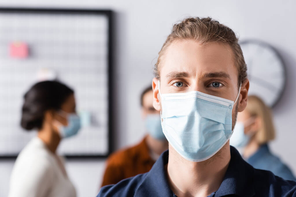 joven gerente de oficina en máscara médica mirando a la cámara cerca de colegas multiculturales en un fondo borroso - Foto, Imagen