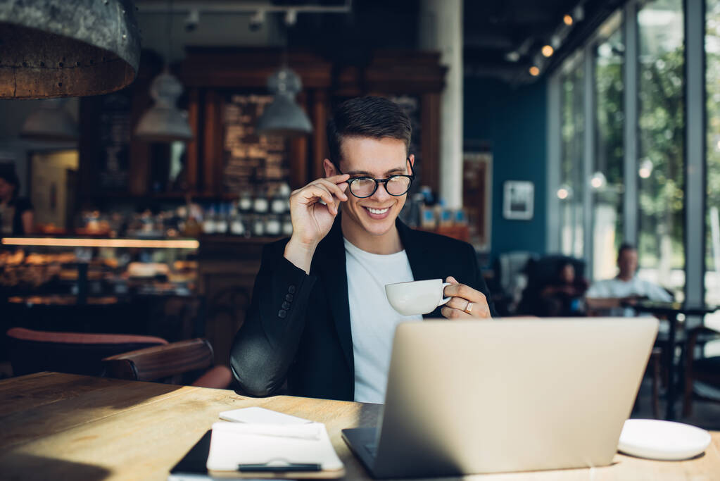 Vrolijke jonge zakenman in bril en formele pak zitten in cafe met kopje koffie en laptop tijdens het werken aan het project - Foto, afbeelding