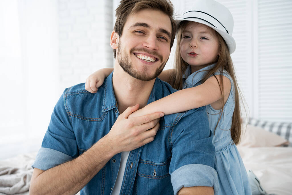 Retrato del padre guapo y su linda hija abrazándose, mirando a la cámara y sonriendo mientras está sentado en el sofá en casa. - Foto, imagen